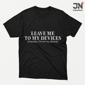 May include: Black t-shirt with the white text "LEAVE ME TO MY DEVICES (Literally. I'm on my phone)". The shirt features a crew neck and short sleeves. The brand logo "JN JokeNest" is in the upper right corner.