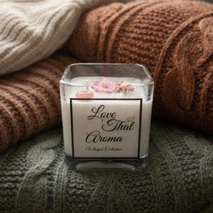 May include: Square glass candle with white wax, topped with a pink flower and a small pink stone. The candle has a black and white label that reads "Love That Aroma" and "Whisper Collection". The candle sits on a knitted surface.