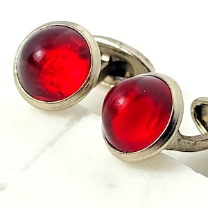 May include: A pair of cufflinks featuring round, translucent red cabochon stones set in polished silver-toned metal frames. The circular red inserts have a smooth, domed surface that reflects light, while the metallic base provides a clean, simple border.