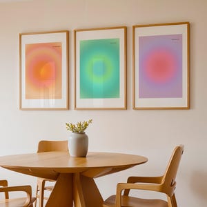 Puede incluir: Tres impresiones art&iacute;sticas enmarcadas con dise&ntilde;os de gradiente coloridos en tonos naranja, verde y morado. Se exhiben sobre una mesa y sillas de madera, con un jarr&oacute;n de flores en el centro. La habitaci&oacute;n tiene una paleta de colores neutros.