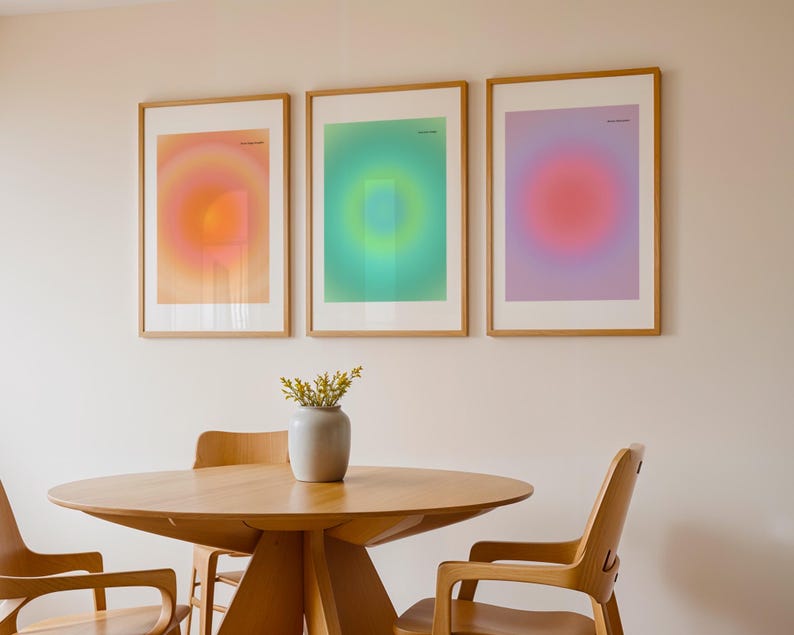 Puede incluir: Tres impresiones art&iacute;sticas enmarcadas con dise&ntilde;os de gradiente coloridos en tonos naranja, verde y morado. Se exhiben sobre una mesa y sillas de madera, con un jarr&oacute;n de flores en el centro. La habitaci&oacute;n tiene una paleta de colores neutros.
