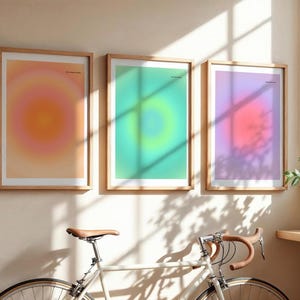 Puede incluir: Tres impresiones art&iacute;sticas enmarcadas con dise&ntilde;os de color degradado en tonos naranja, verde y morado. Las impresiones est&aacute;n en marcos de madera y cuelgan de una pared beige. Una bicicleta con sill&iacute;n y manillar marrones est&aacute; en primer plano.