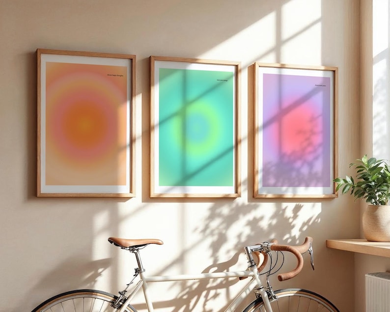 Puede incluir: Tres impresiones art&iacute;sticas enmarcadas con dise&ntilde;os de color degradado en tonos naranja, verde y morado. Las impresiones est&aacute;n en marcos de madera y cuelgan de una pared beige. Una bicicleta con sill&iacute;n y manillar marrones est&aacute; en primer plano.
