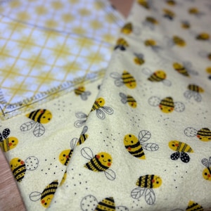 May include: Two soft, folded cloths. One cloth features a yellow and white checkered pattern. The other cloth has a light yellow background with a repeating pattern of smiling cartoon bees with black and yellow stripes and gray wings.