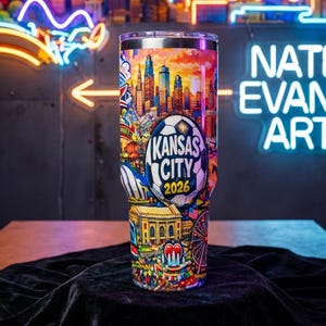 May include: A stainless steel tumbler with a colorful design featuring a cityscape, a soccer ball, and the text "Kansas City 2026." The design includes a Ferris wheel, a heart-shaped fountain, and other illustrations. The tumbler is cylindrical and has a clear lid.