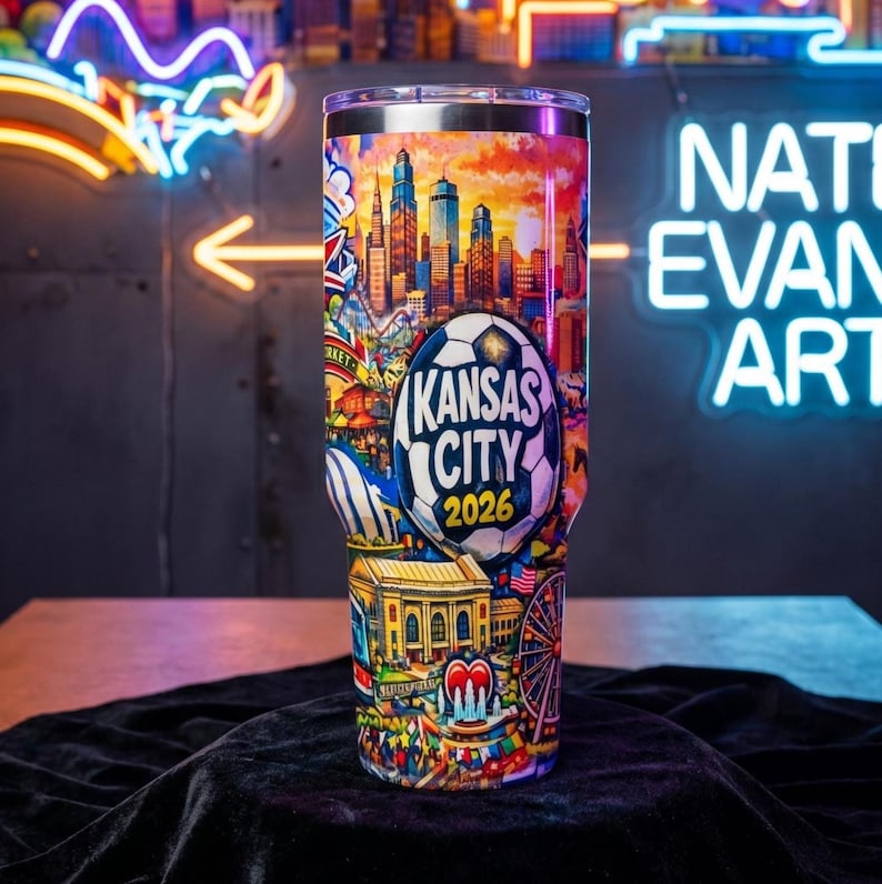 May include: A stainless steel tumbler with a colorful design featuring a cityscape, a soccer ball, and the text "Kansas City 2026." The design includes a Ferris wheel, a heart-shaped fountain, and other illustrations. The tumbler is cylindrical and has a clear lid.