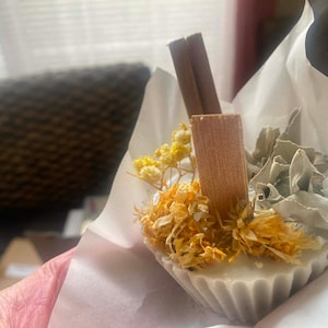 Könnte beinhalten: Eine weiße, cupcake-förmige Kerze mit getrockneten Blumen, Salbei und Zimtstangen. Die Kerze hat einen geriffelten Rand und ist in weißes Papier gewickelt. Die getrockneten Blumen sind gelb und orange.