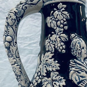 May include: A dark blue ceramic pitcher with a white handle and rim. The pitcher is decorated with white grape vines, leaves, and figures. The base is a light beige color.
