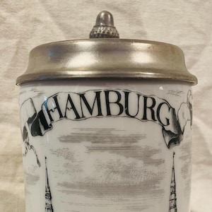 May include: A white ceramic beer stein with a metallic lid featuring a black and white illustration of the Hamburg city skyline. The word HAMBURG is printed in a banner at the top, with detailed depictions of church spires, buildings, and boats.