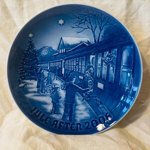 May include: A round blue and white porcelain decorative plate featuring a winter scene of people boarding a train at a station with a Christmas tree. The text JULE AFTEN 2006 is printed in a dark blue font along the bottom edge of the plate.