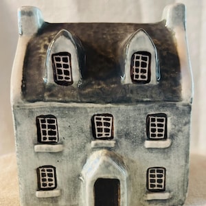 Könnte beinhalten: Keramikhaus-Figur mit grauem und braunem Dach, hellblauen Wänden und schwarzen Fenstern. Das Haus hat eine kleine Eingangstür und Dachgauben. Das Design erinnert an ein gemütliches Cottage.