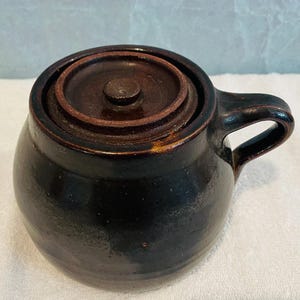 Może przedstawiać: Ciemnobrązowy ceramiczny garnek z pokrywką i uchwytem. Garnek ma zaokrąglony korpus i pokrywkę, która idealnie pasuje na górę. Uchwyt jest łukowaty i przymocowany do boku. Ceramika ma błyszczące wykończenie.