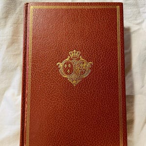 Puede incluir: Un libro de tapa dura con una cubierta marrón rojiza texturizada y detalles dorados. La cubierta presenta un emblema dorado con una corona y un diseño ornamentado. Los bordes del libro también están adornados en dorado.