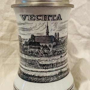 以下が含まれることがあります： 金属製の蓋が付いた白い陶器製のビールジョグ。フェヒタの町の黒いインクのイラストが描かれています。デザインには、建物、教会、風景、そして容器の底の周りにある装飾的な花柄が含まれています。