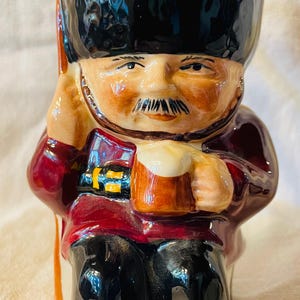 Puede incluir: Figura de cerámica de un guardia real británico, con sombrero de piel de oso negro, túnica roja y pantalón negro. La figura sostiene una jarra de cerveza y un bastón. Los detalles están pintados a mano con un acabado brillante.