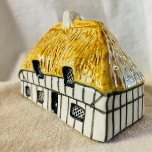 May include: A ceramic cottage figurine with a thatched roof. The roof is a light yellow color, and the walls are white with black vertical stripes. The windows are black with a crosshatch pattern. A small chimney is on the roof.