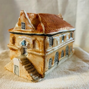 May include: A ceramic house-shaped trinket box with a detailed design. The roof is a reddish-brown colour, and the walls are a light tan with white trim. The windows have a grid pattern. A small staircase leads to a door.