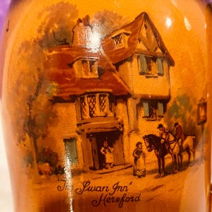 Puede incluir: Jarra de cerámica con una pintura detallada de "The Swan Inn" en Hereford. La obra de arte representa un edificio de dos pisos con un tejado rojo, figuras y caballos. La jarra tiene una cálida combinación de colores naranja y marrón.