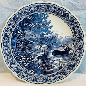 Puede incluir: Un plato decorativo redondo, blanco y azul, con borde festoneado. El plato presenta una escena de paisaje azul con un ciervo en un cuerpo de agua, rodeado de árboles y follaje. El borde tiene un diseño floral y de volutas.