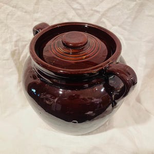 May include: A dark brown ceramic bean pot with a glossy finish, featuring a rounded body, a matching lid with a central knob, and a sturdy side handle. The pot has a smooth, reflective surface and a wide opening, showcasing a classic, functional design.
