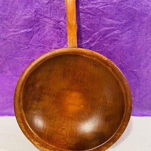May include: A round, wooden bowl with a long handle. The bowl is a warm brown color with visible wood grain. The handle is attached to the bowl with a small metal ring. The bowl is likely used for serving or display.