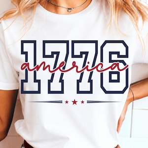 May include: A white short-sleeved t-shirt featuring the navy blue block-style text 1776 with the word america written in a red cursive script across the center. Below the text are three small red stars centered between two horizontal navy lines.