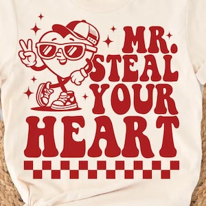 May include: Cream-colored t-shirt with a red graphic of a heart character wearing sunglasses and a cap, giving a peace sign. The text "MR. STEAL YOUR HEART" is in red, with a checkered pattern at the bottom.