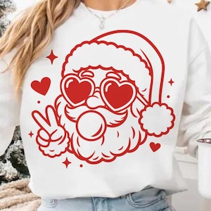 May include: White sweatshirt with a red Santa Claus graphic wearing heart-shaped sunglasses and giving a peace sign. A white mug with a Santa Claus graphic is held in the hand. Christmas tree and decorations are in the background.