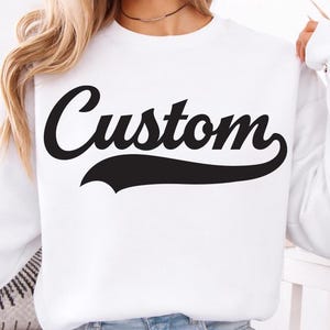 May include: White sweatshirt with the word "Custom" in a black, cursive font. The word is underlined with a black swoosh. The sweatshirt is long-sleeved and the model is wearing light blue denim shorts.