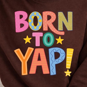 May include: A brown sweatshirt with the words "BORN TO YAP!" in colorful, playful lettering. The text features various patterns and star accents, creating a fun and eye-catching design. The sweatshirt is a casual, comfortable style.