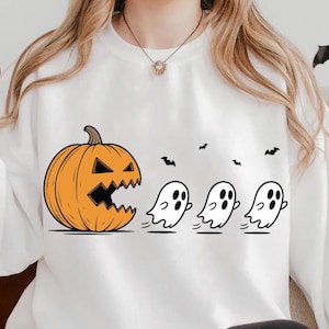 May include: A white long-sleeve sweatshirt featuring a graphic of a large orange jack-o'-lantern chasing three small white ghosts. Small black bats are scattered above the ghosts. A black mug with the text Witches Brew is held in one hand.