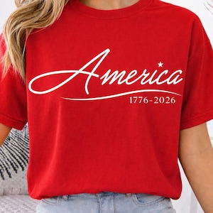 May include: A red t-shirt with the word "America" in white script, accompanied by a star and the dates "1776-2026". The shirt has short sleeves and a crew neck.