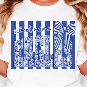 May include: A white t-shirt featuring a blue and white striped background graphic with the text school name and EAGLES in a blue leopard print pattern. A large blue and white striped bow is positioned on the right side of the design.