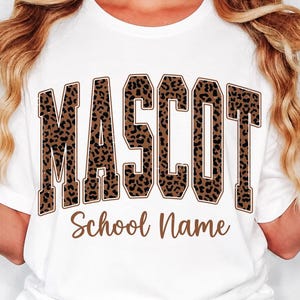 Può includere: Una t-shirt bianca a maniche corte con la parola MASCOT in grandi lettere in stile blocco con un motivo leopardato marrone e nero. Sotto la parola MASCOT, il testo School Name è stampato in un carattere corsivo di colore marrone.