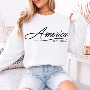 May include: White sweatshirt with the word "America" in a cursive font, with a star replacing the letter "i". Below the word is the text "1776-2026". The sweatshirt has long sleeves.