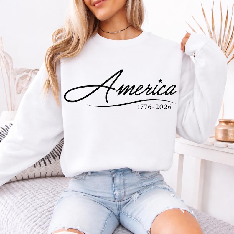 May include: White sweatshirt with the word "America" in a cursive font, with a star replacing the letter "i". Below the word is the text "1776-2026". The sweatshirt has long sleeves.