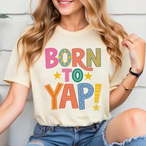 May include: Cream-colored t-shirt with the text "BORN TO YAP!" in colorful, playful lettering. The design includes pink, orange, green, yellow, and blue elements, with star accents. The shirt is a casual, short-sleeved style.
