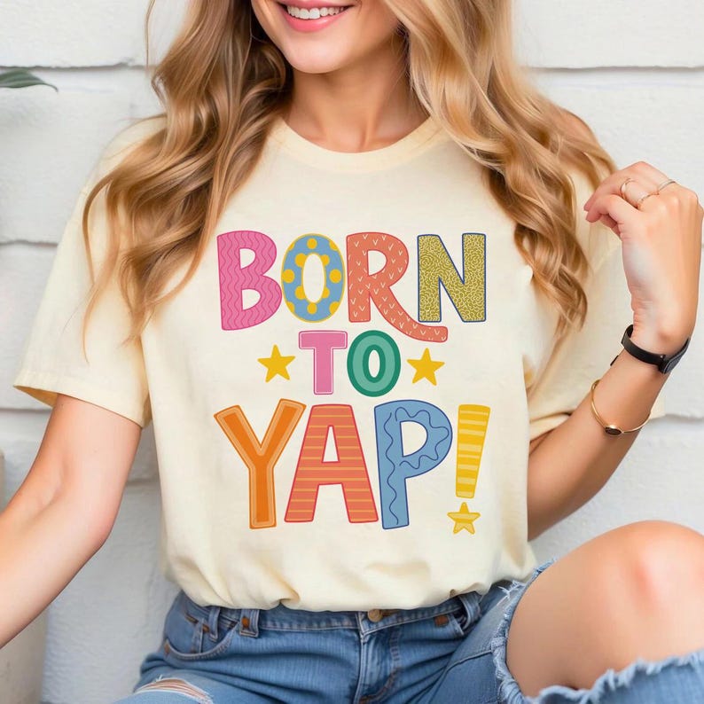May include: Cream-colored t-shirt with the text "BORN TO YAP!" in colorful, playful lettering. The design includes pink, orange, green, yellow, and blue elements, with star accents. The shirt is a casual, short-sleeved style.