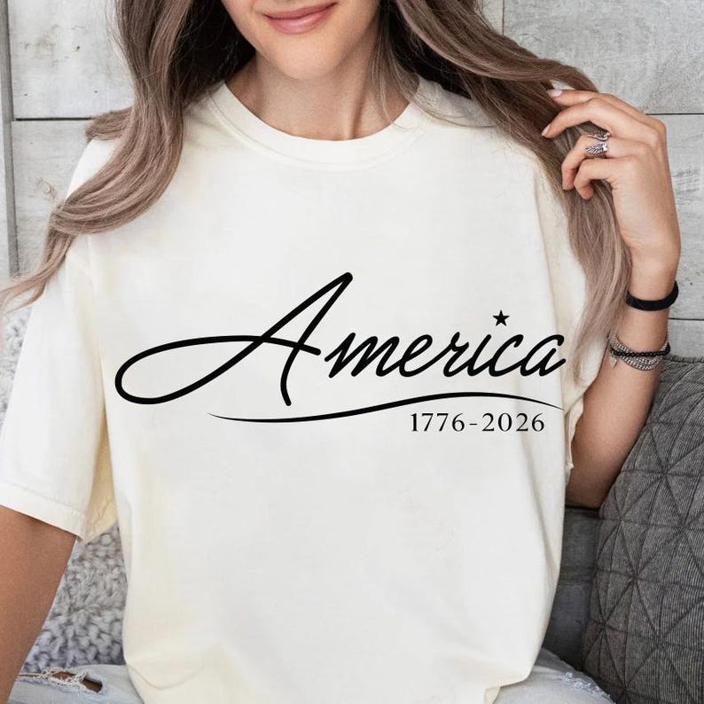 May include: Cream-colored t-shirt with the word "America" in a black script font. Below the word is the text "1776-2026". The shirt is a casual style, suitable for everyday wear.