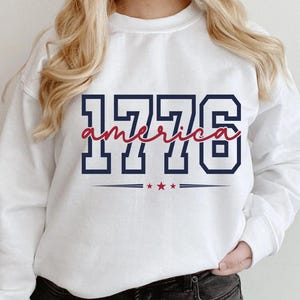 May include: A white long-sleeve sweatshirt featuring the navy blue block-style text 1776 with the word america written in red cursive script across the center. Below the text are three small red stars centered between two horizontal navy blue lines.