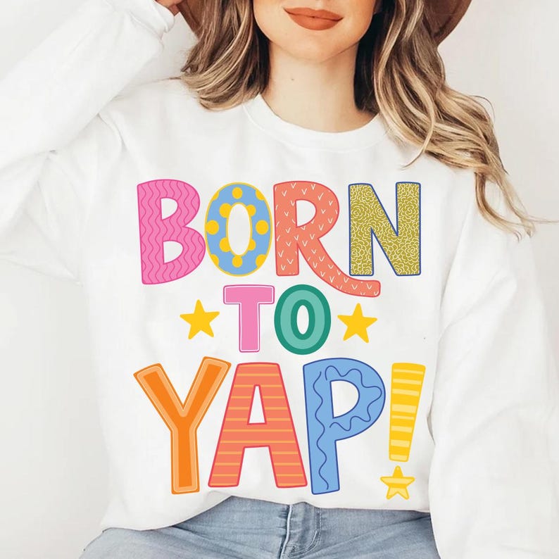 May include: White sweatshirt with the phrase "BORN TO YAP!" in colorful, playful lettering. The text features various patterns and colors, including pink, blue, yellow, orange, and green. Two yellow stars are also included in the design.