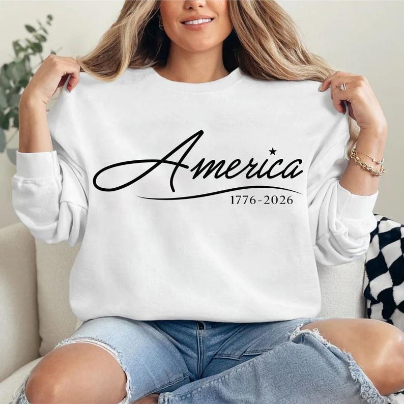 May include: White sweatshirt with the word "America" in a cursive font, with a star and the dates "1776-2026" printed below. The sweatshirt has long sleeves and is worn with blue jeans.