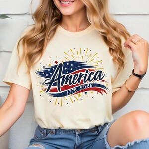 May include: A cream-coloured t-shirt with the word "America" in a decorative font, set against a red and blue banner design. The design incorporates stars, fireworks, and the dates "1776-2026".
