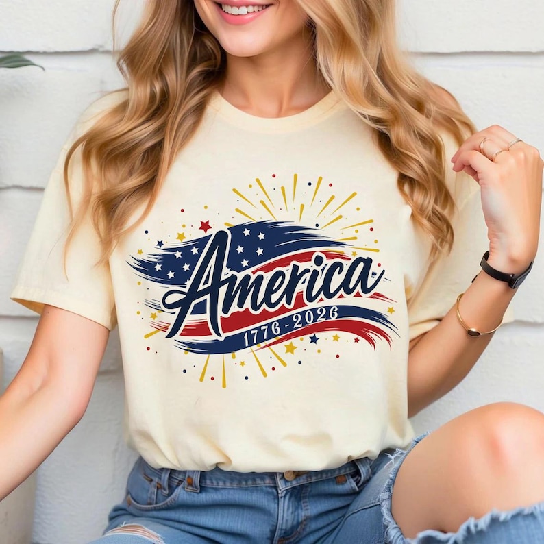 May include: A cream-coloured t-shirt with the word "America" in a decorative font, set against a red and blue banner design. The design incorporates stars, fireworks, and the dates "1776-2026".
