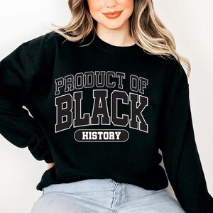 May include: Black sweatshirt with the text "PRODUCT OF BLACK HISTORY" in white block letters. The sweatshirt is a crew neck style and appears to be made of a soft material. The text is outlined in black.