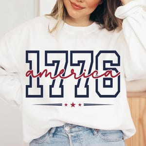 May include: A white long-sleeve sweatshirt featuring the navy blue block-style text 1776 with the word america written in red cursive script across the center. Below the text are three small red stars flanked by two horizontal navy blue lines.