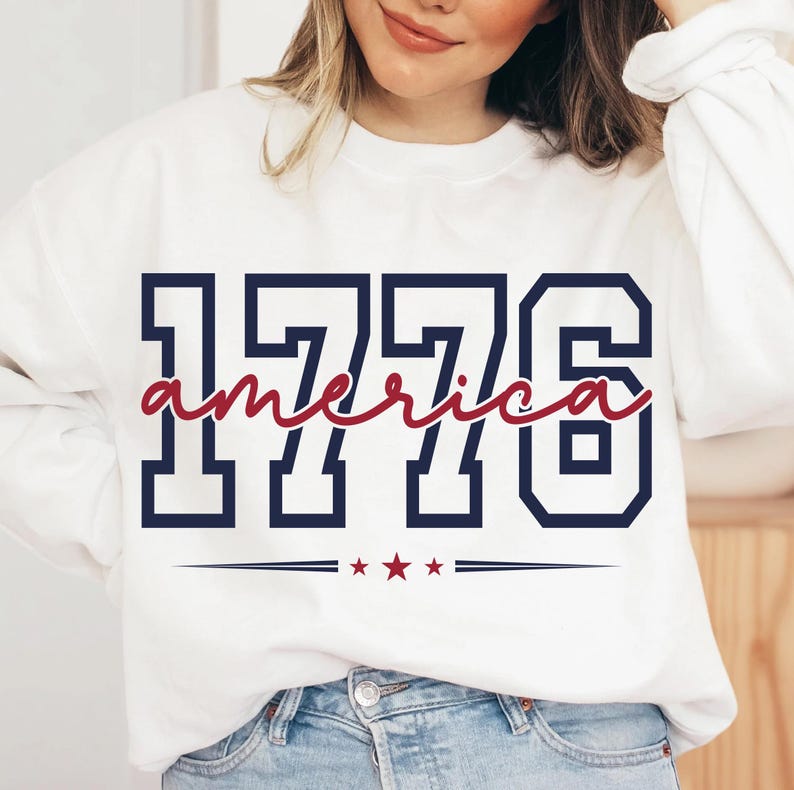 May include: A white long-sleeve sweatshirt featuring the navy blue block-style text 1776 with the word america written in red cursive script across the center. Below the text are three small red stars flanked by two horizontal navy blue lines.