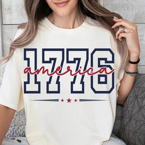 May include: A cream-colored short-sleeve t-shirt featuring the navy blue block-style numbers 1776. The word america is written in a red cursive script across the center of the numbers, with three small red stars centered below the text.