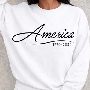 May include: White sweatshirt with the word "America" in a cursive font, with a star above the "i". Below the word is the text "1776-2026". The sweatshirt is a solid white color.