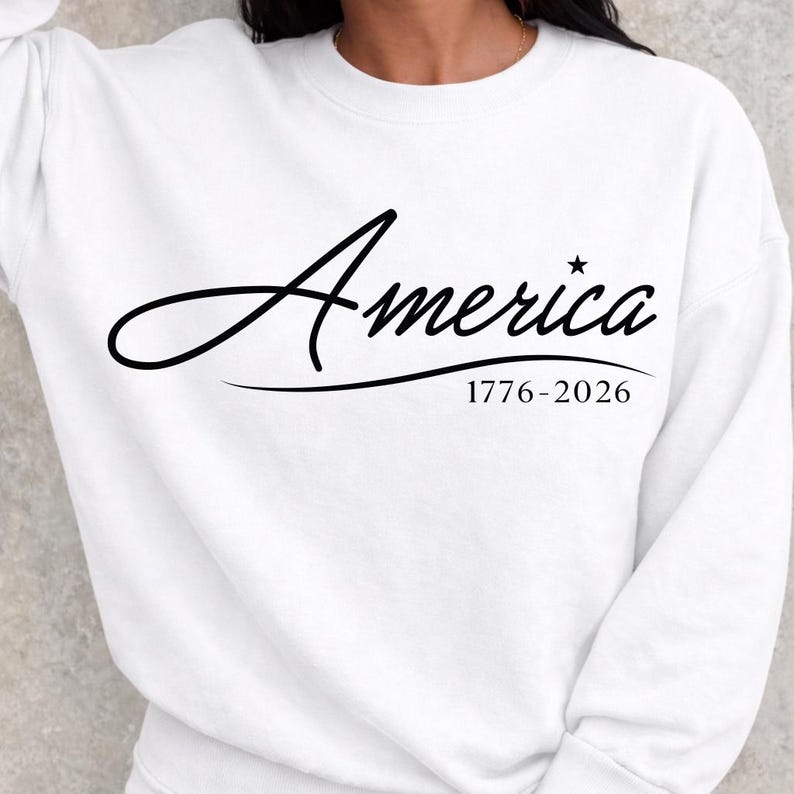 May include: White sweatshirt with the word "America" in a cursive font, with a star above the "i". Below the word is the text "1776-2026". The sweatshirt is a solid white color.